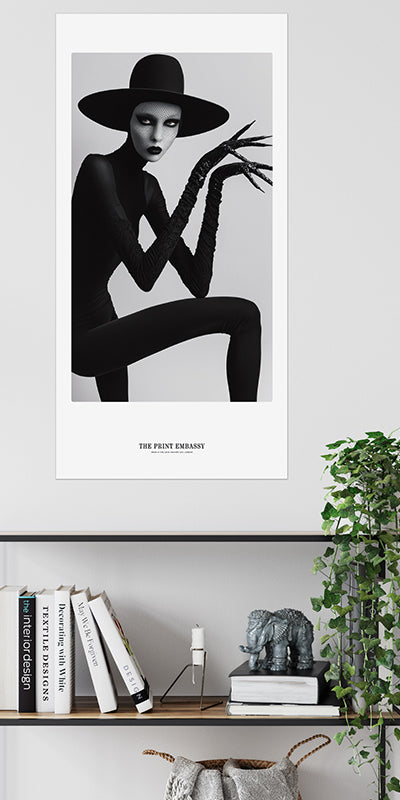 A statement black and white poster of a person in a black outfit and large Boy George styled hat, the model is wearing long, spooky clawed gloves. It could be a still from a Marilyn Manson music video, hanging above a modern black shelf.