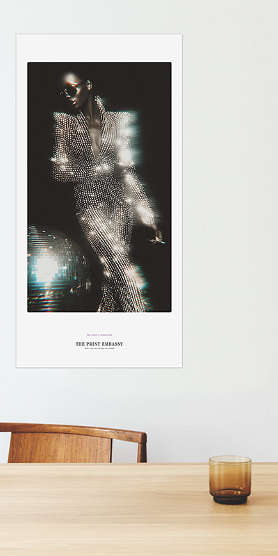 Poster of a person in a sparkling outfit next to a disco ball, the oversized poster is on a wall above a table with a glass.