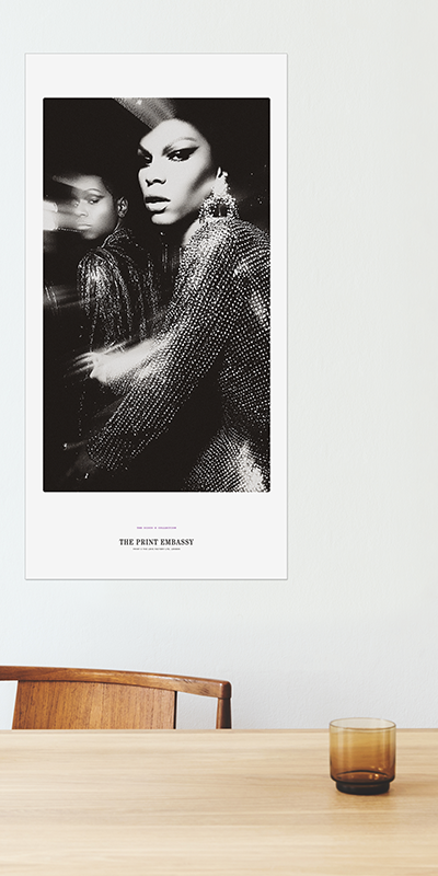 Poster of a gorgeous drag queen in a black and white setting on a wall above a wooden table. It could be from the cover of any Vogue.